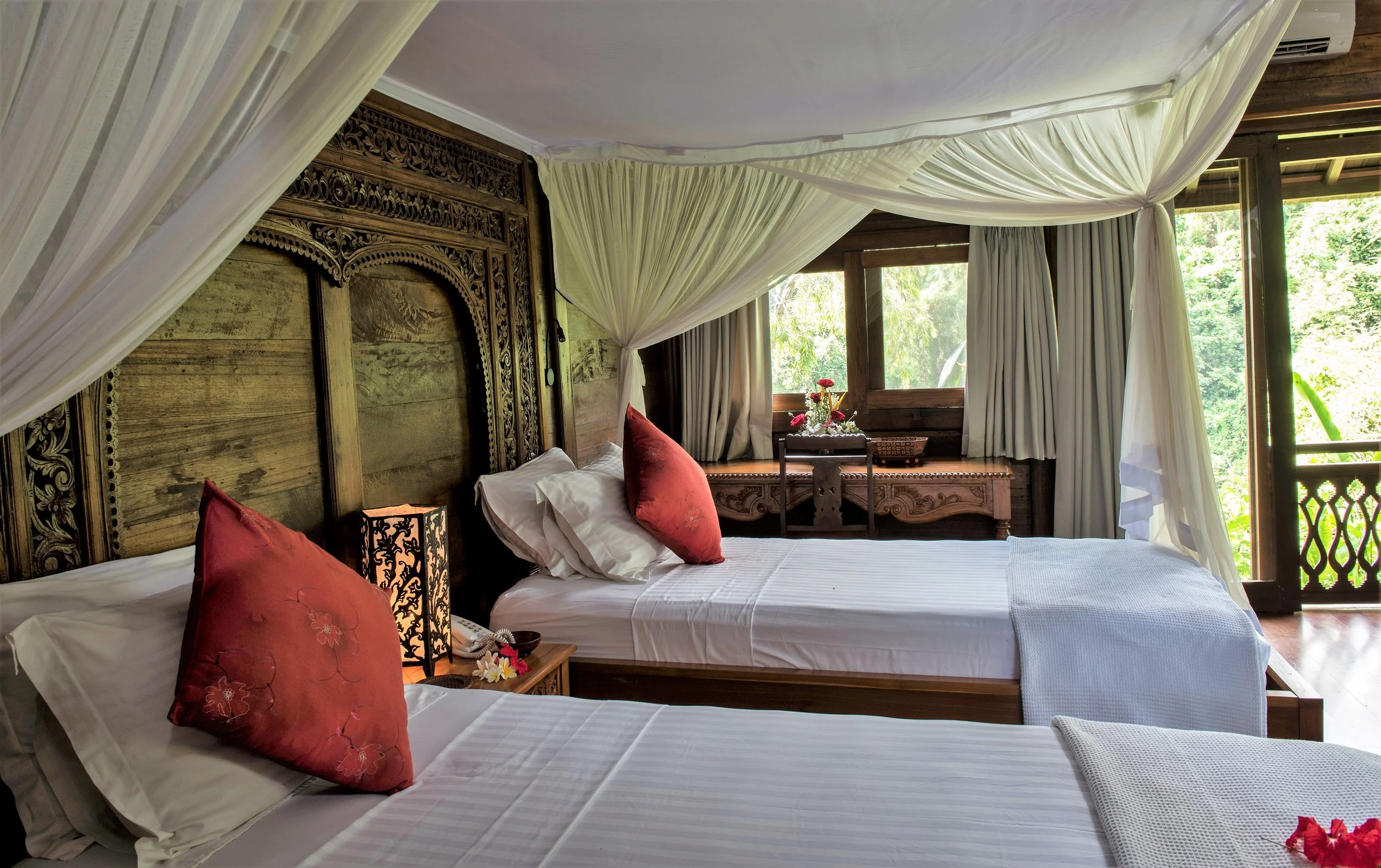 A bedroom with two single beds, red pillows, wooden headboards, a bedside lamp, and a window with curtains overlooking greenery.
