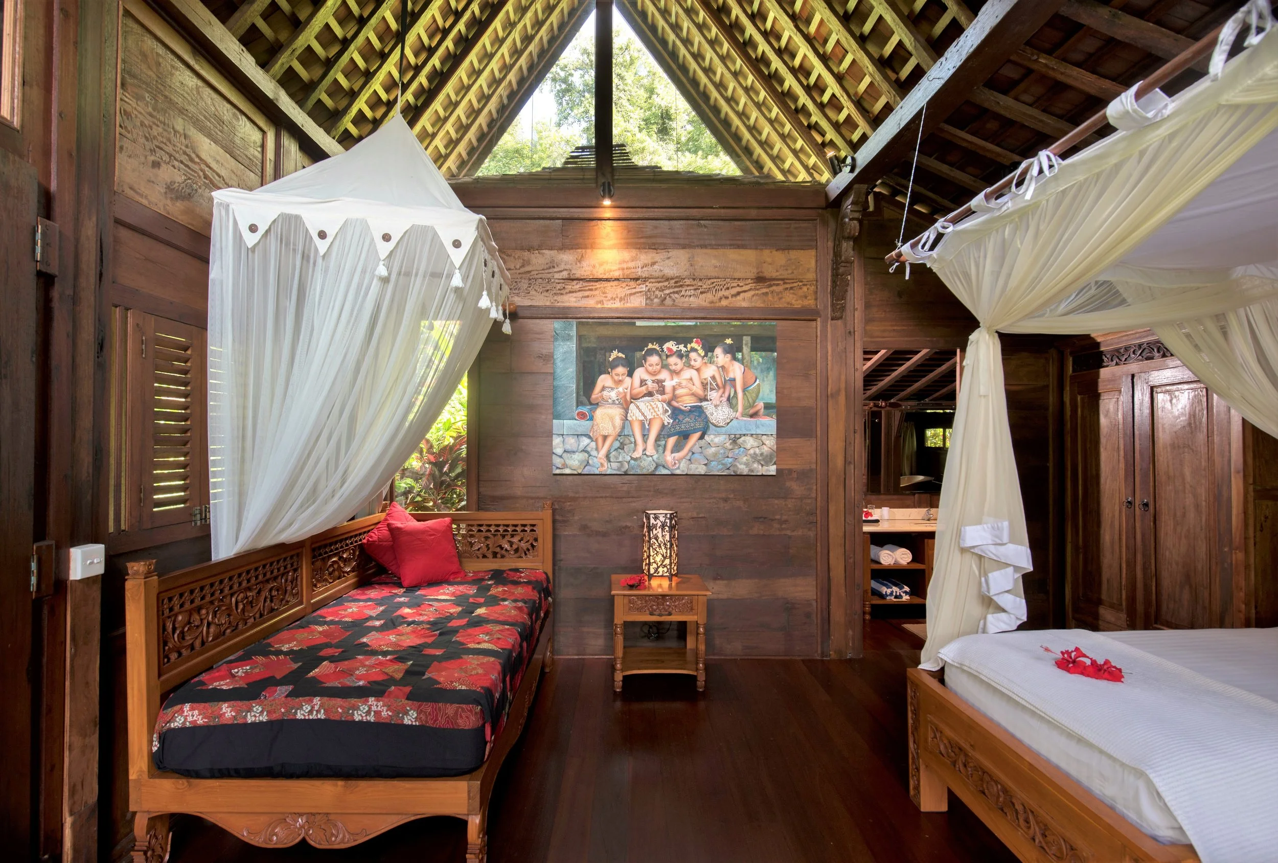 A cozy bedroom with wooden walls and ceiling, two beds with white and red bedspreads, a small wooden nightstand with a decorative lamp, a painting of children in traditional attire on the wall, and a window with white curtains and greenery outside.