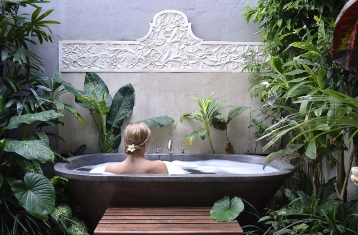 A woman with blonde hair, wearing a white flower in her hair, is relaxing in an outdoor bathtub surrounded by lush green tropical plants. The bath area features a stone wall with decorative carvings at the top.