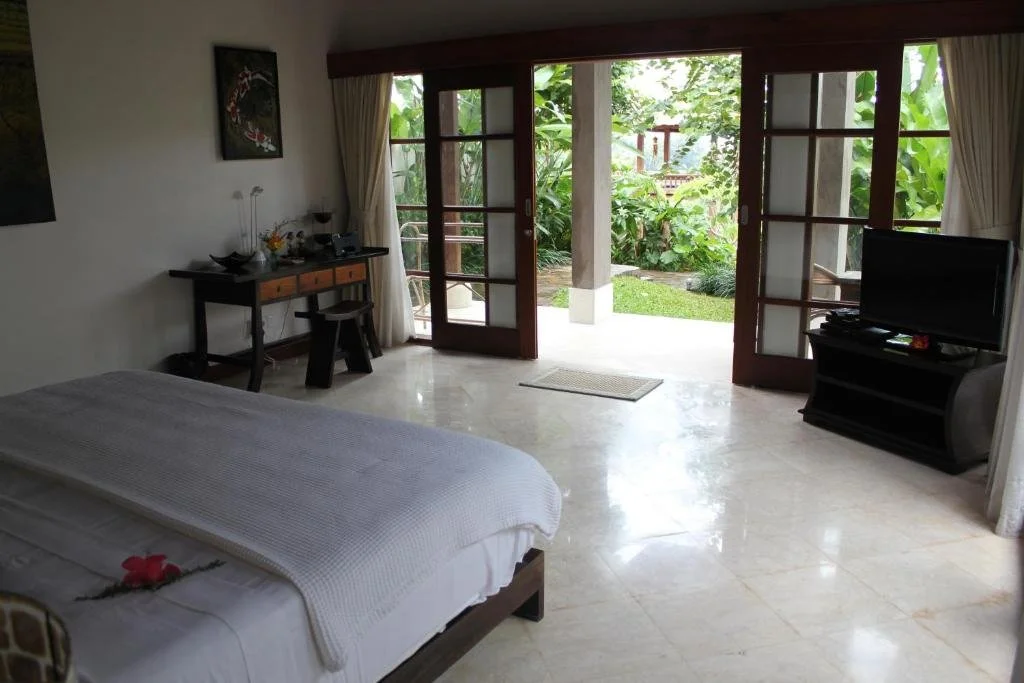 A bedroom with a bed, a desk, a TV, and large glass doors leading to a garden with lush greenery.