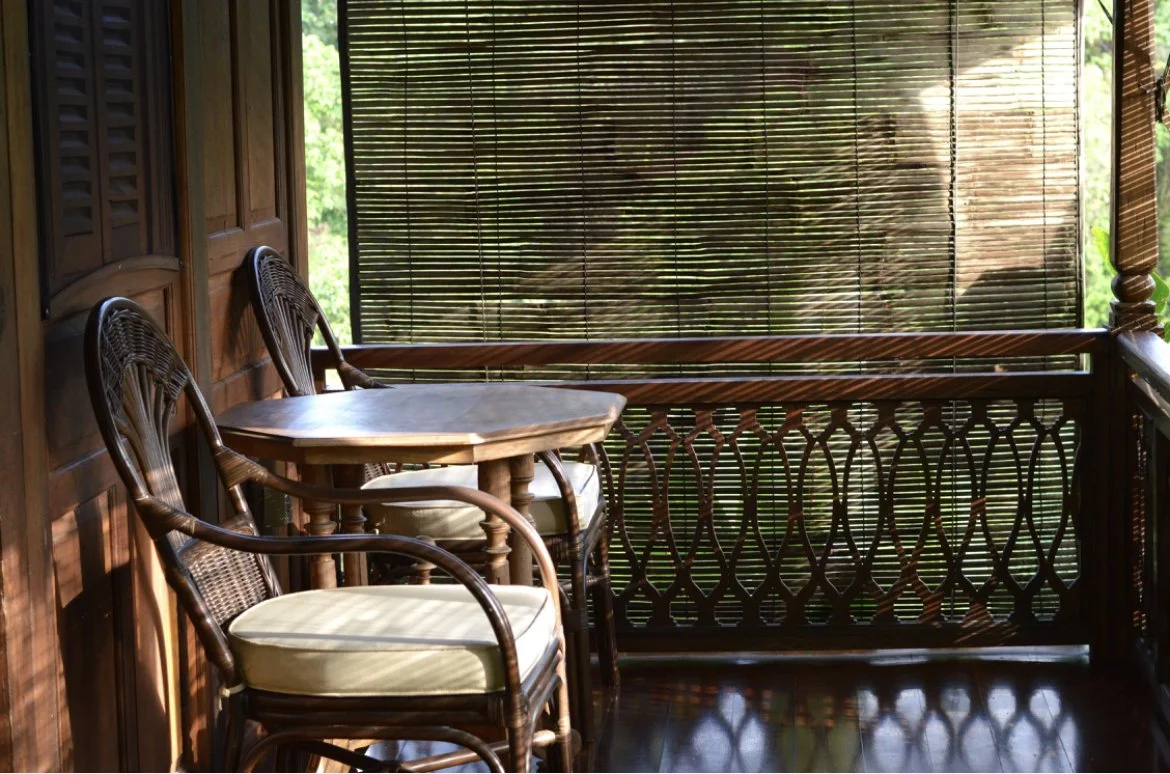 A small balcony with two wooden chairs with cushioned seats and a round table, surrounded by bamboo blinds and wooden railing, overlooking green trees outside.