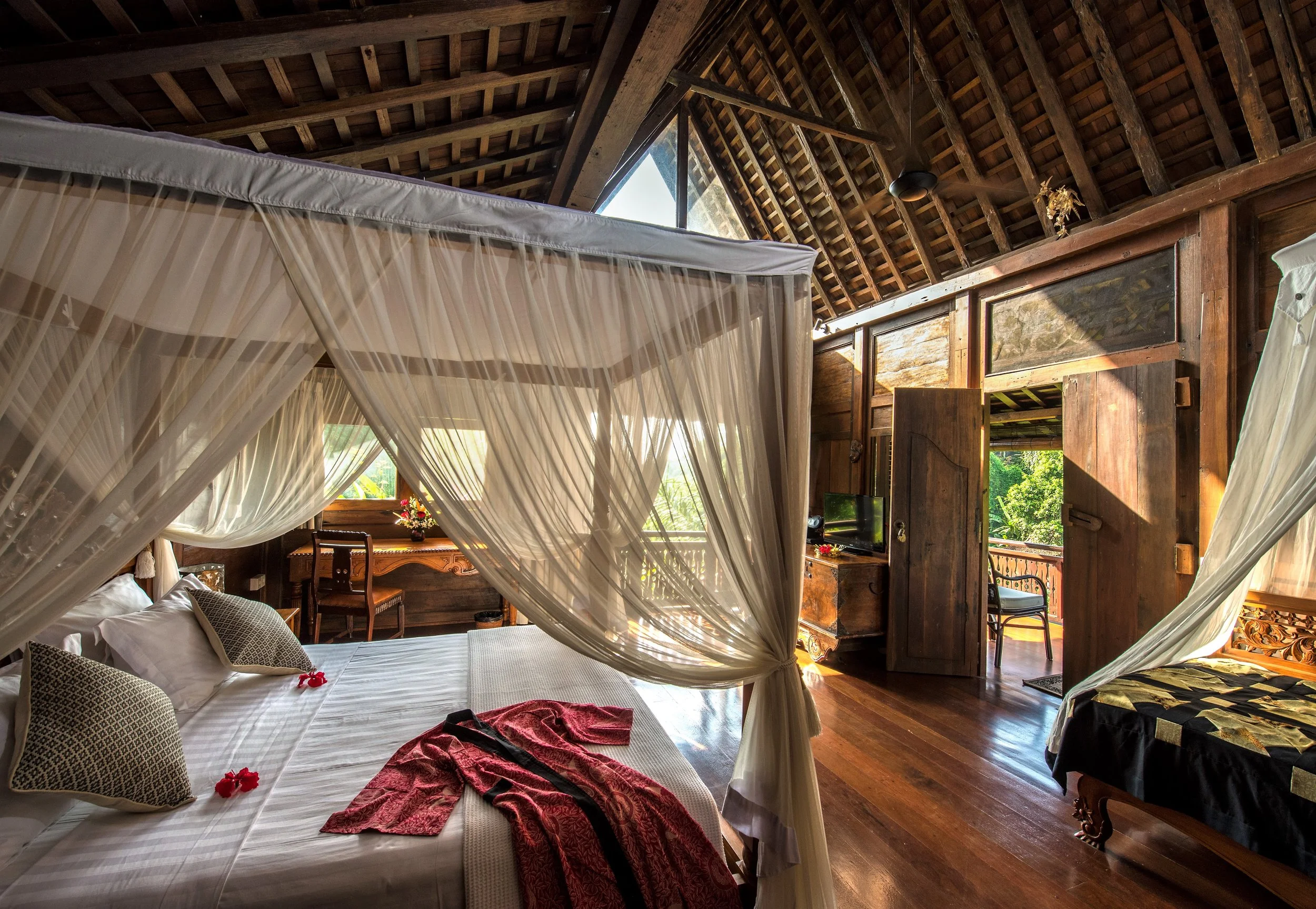Interior of a rustic wooden bedroom with a four-poster bed and a mosquito net, with natural light streaming through windows and an open balcony door showing greenery outside.