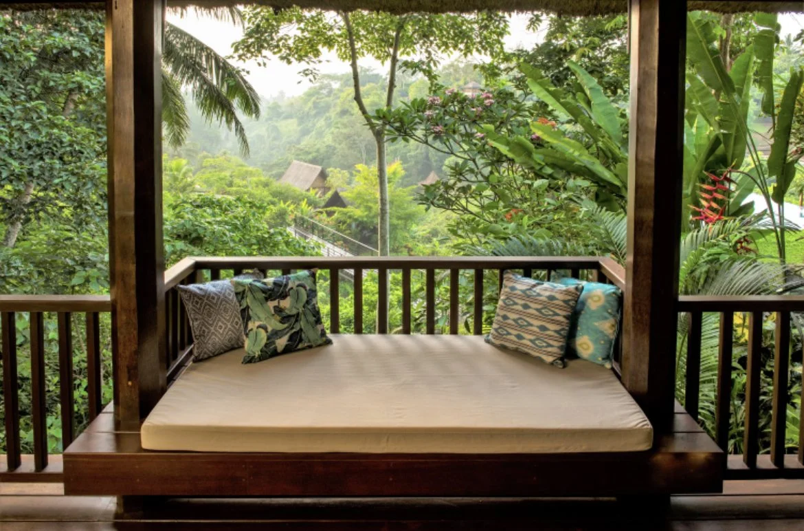Wooden outdoor balcony with a mattress and pillows overlooking a lush, green jungle landscape.