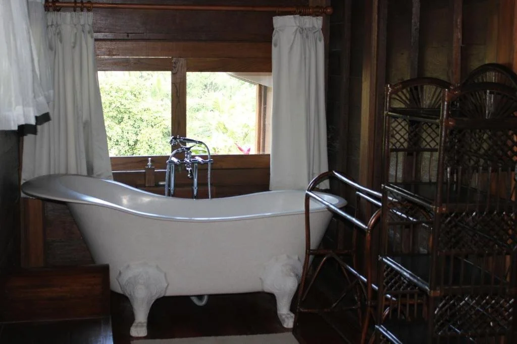 Vintage white clawfoot bathtub inside a rustic wooden bathroom with a window, curtains, and dark wooden shelving.