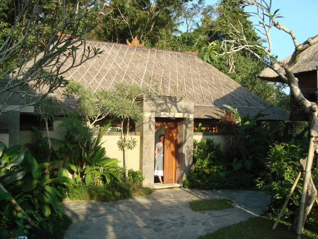 A woman standing in the doorway of a tropical cottage surrounded by lush greenery and trees, with a stone pathway leading up to the door.