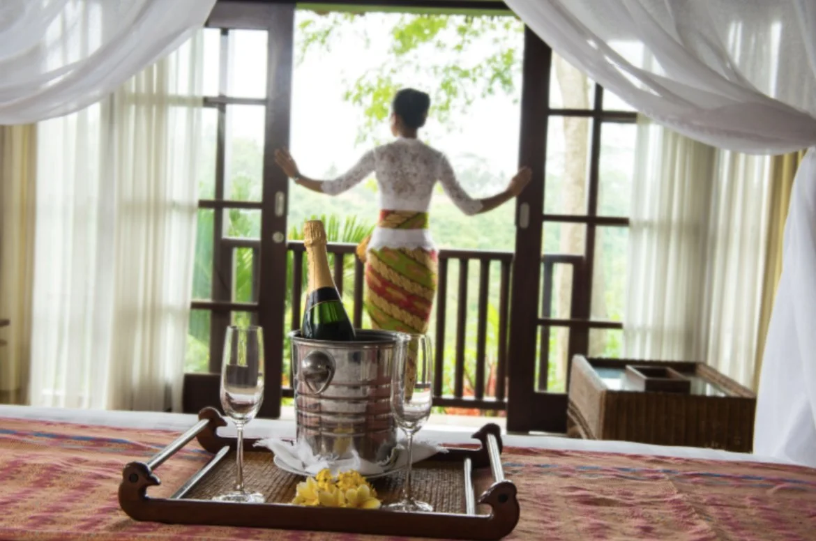 A woman in traditional dress looking out from a wooden balcony over lush green trees, with a table set for celebration including a champagne bottle in an ice bucket, two glasses, and floral decorations.