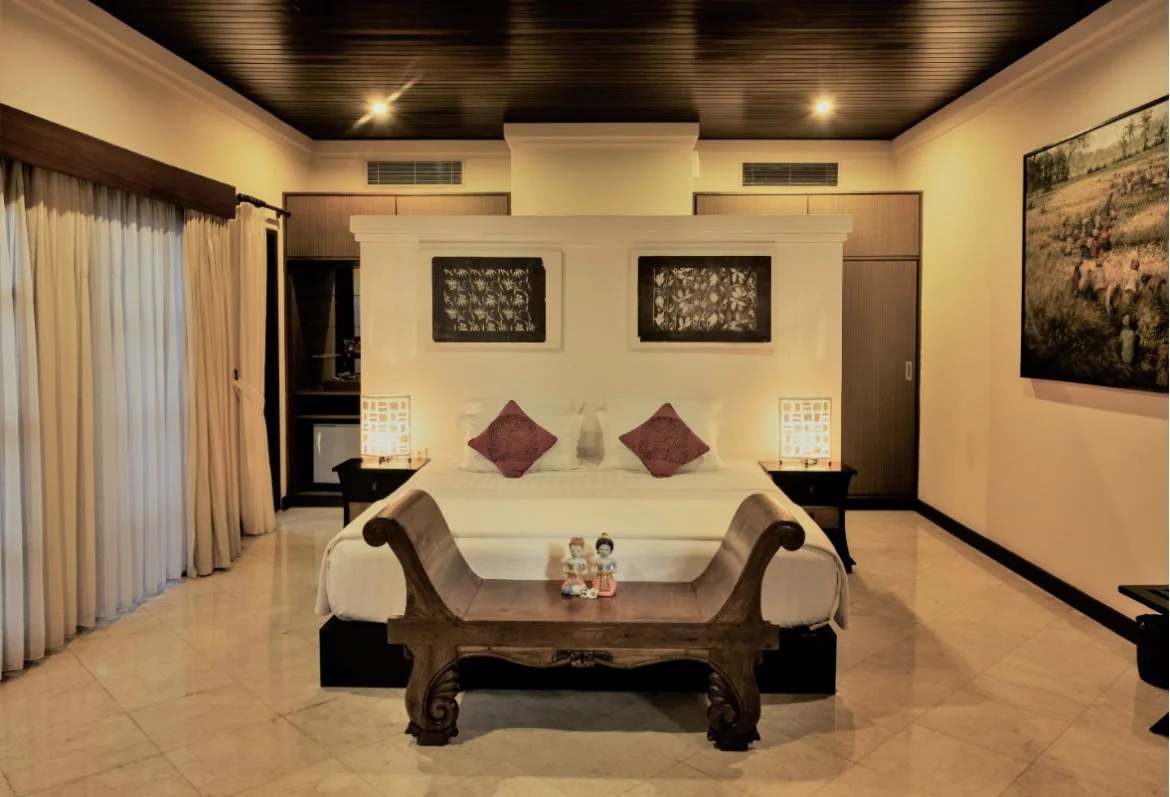 A hotel room with a large bed, two side tables with lamps, a wooden bench at the foot of the bed, and artwork on the walls, featuring cream-colored walls, a dark wooden ceiling, and beige tiled flooring.