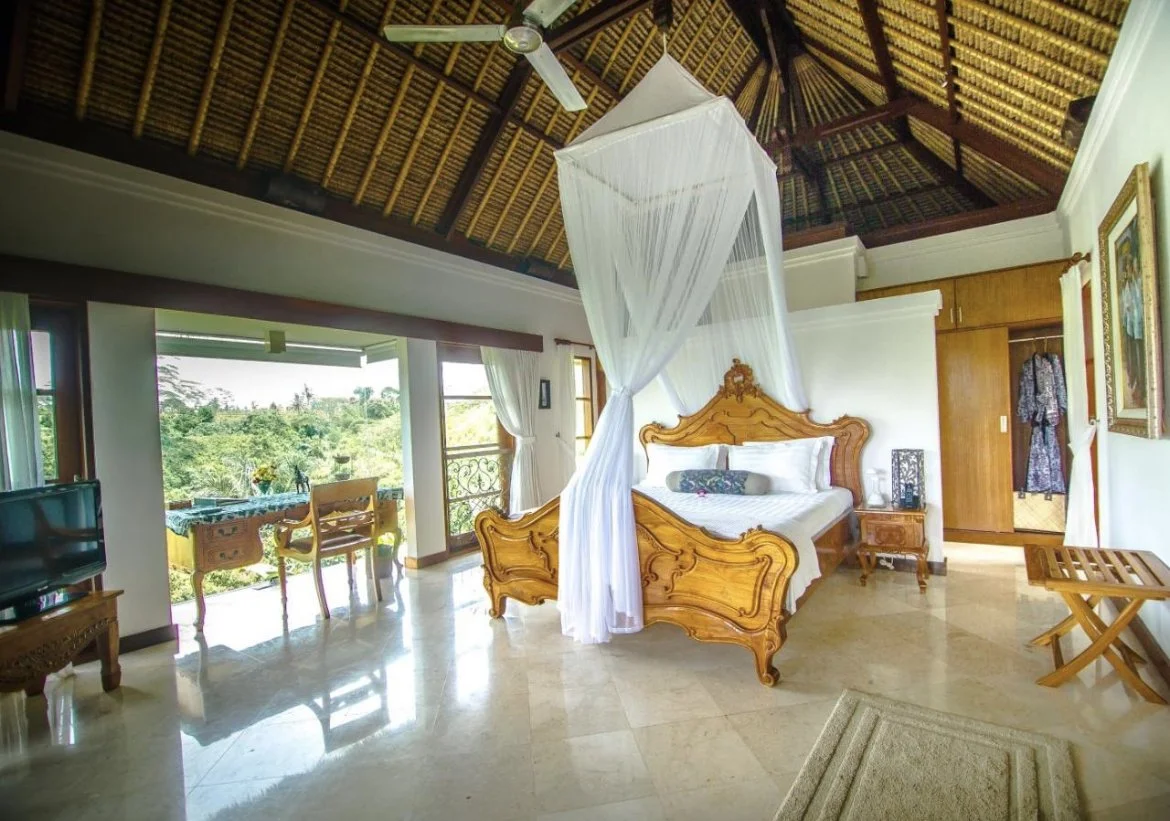 Hotel bedroom with a wooden bed and white mosquito net, sliding glass doors opening to lush greenery, and wooden ceiling with thatch design.