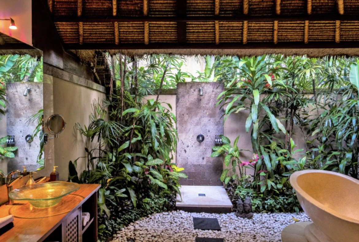 Outdoor bathroom with lush tropical plants, stone shower, wooden sink counter, glass vessel sink, and a bathtub, featuring natural and tropical decor.