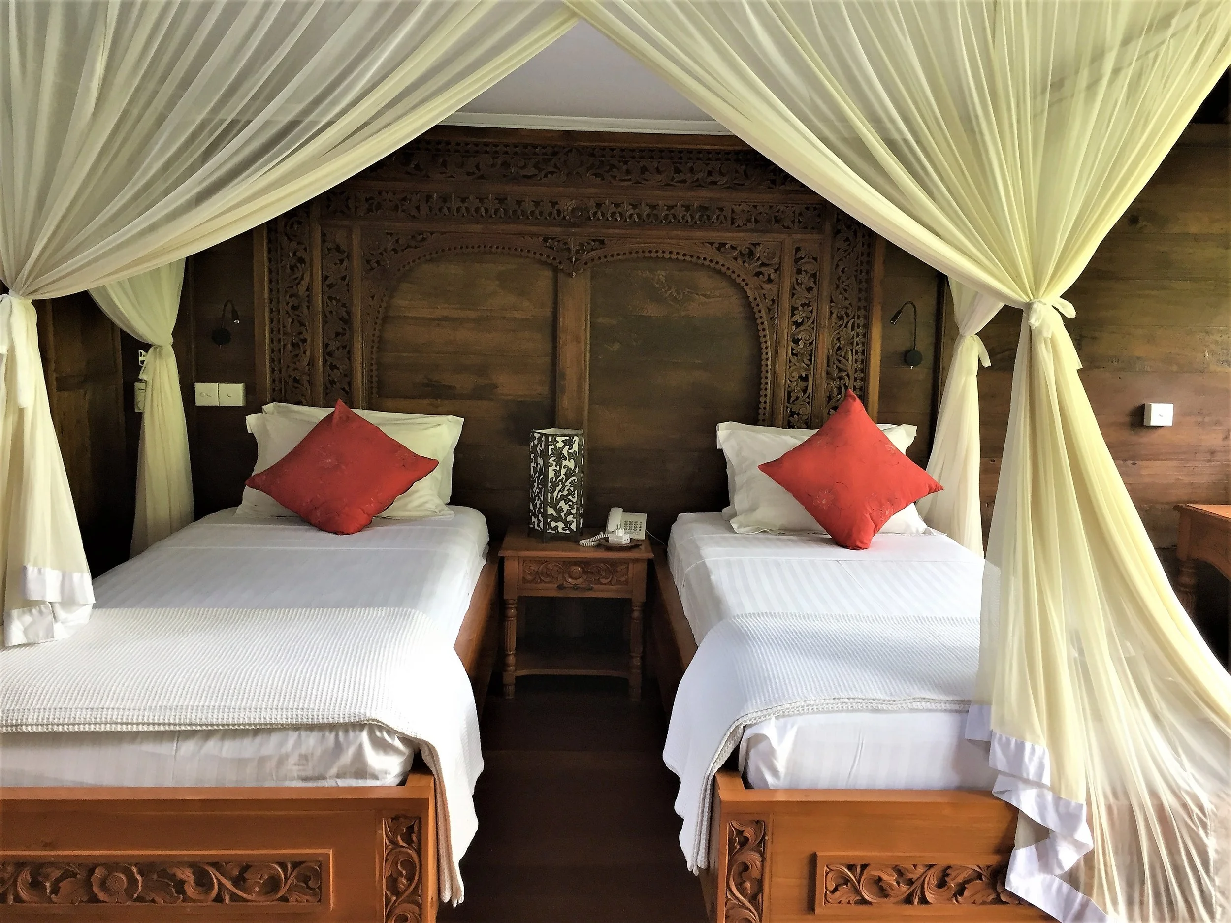 Two beds with white linens and decorative red pillows, framed by cream-colored canopy curtains, featuring a dark wooden headboard with detailed carvings, a bedside table with a lamp and a telephone, in a cozy hotel room.