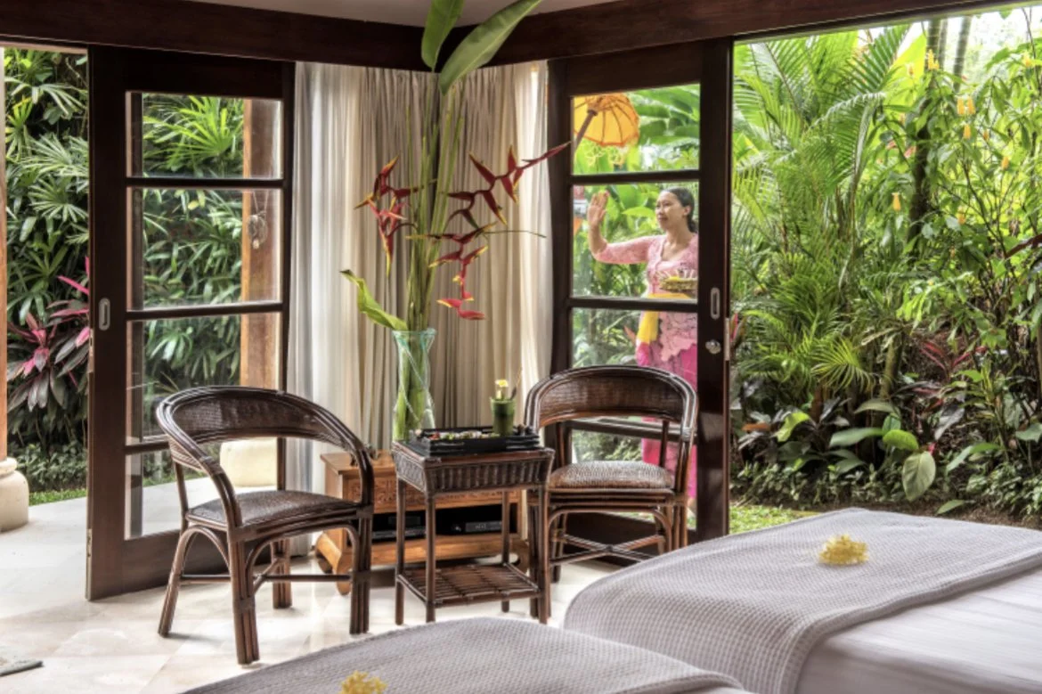 A bedroom with an open sliding glass door revealing a lush tropical garden. A woman in traditional attire stands outside, waving from behind the door. Inside, there are two wicker chairs, a table with a flower arrangement, and tropical plants visible