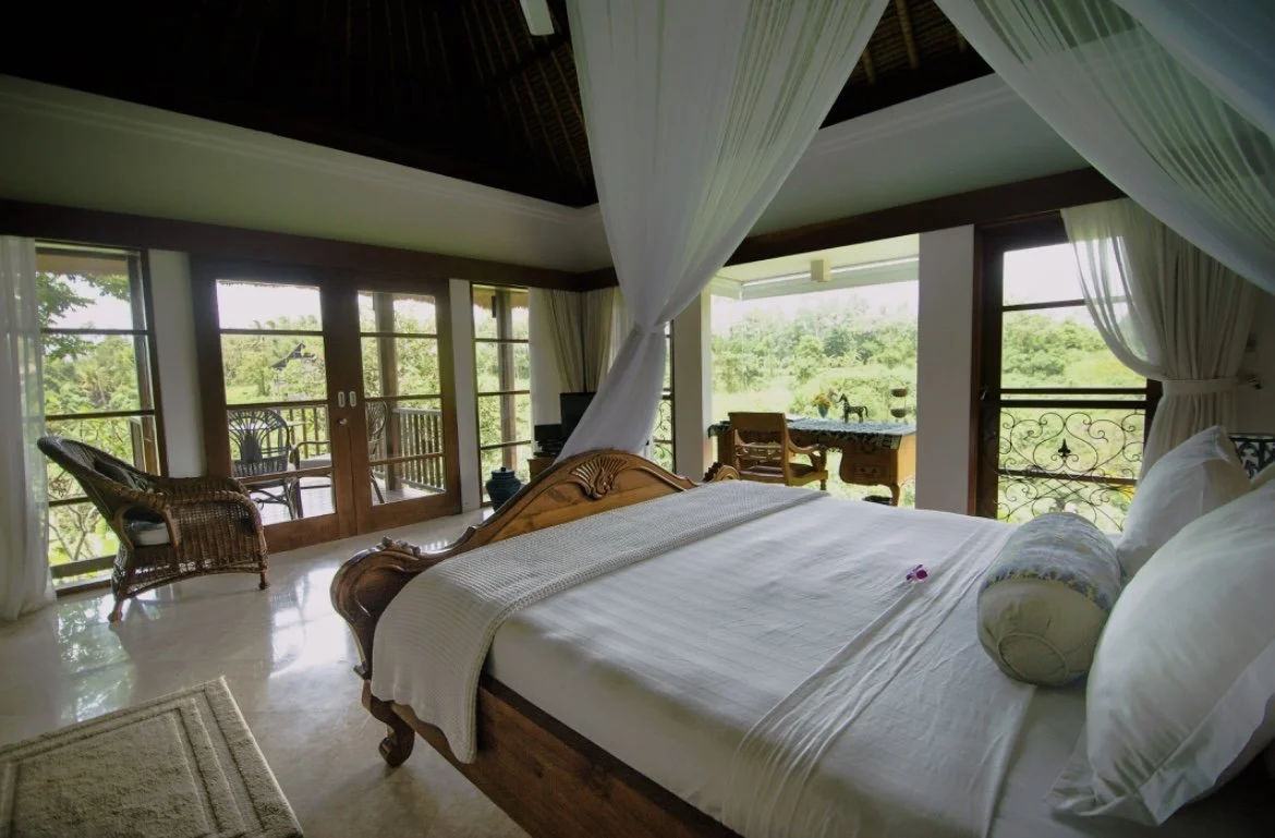 A cozy bedroom with large windows and a balcony, featuring a wooden bed, white bedding, and sheer curtains, overlooking lush greenery outside.