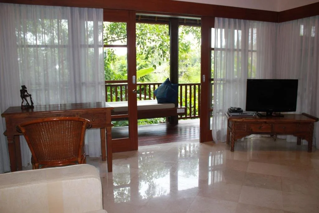 Living room with sliding glass doors leading to a balcony with greenery, wooden furniture including a TV stand, desk, and outdoor bench with a pillow on it, and sheer white curtains.