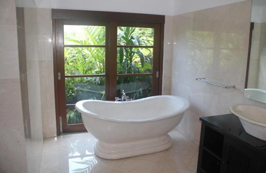 Bathroom with a freestanding bathtub in front of a large window showing lush greenery outside, and a black vanity with a vessel sink to the right.
