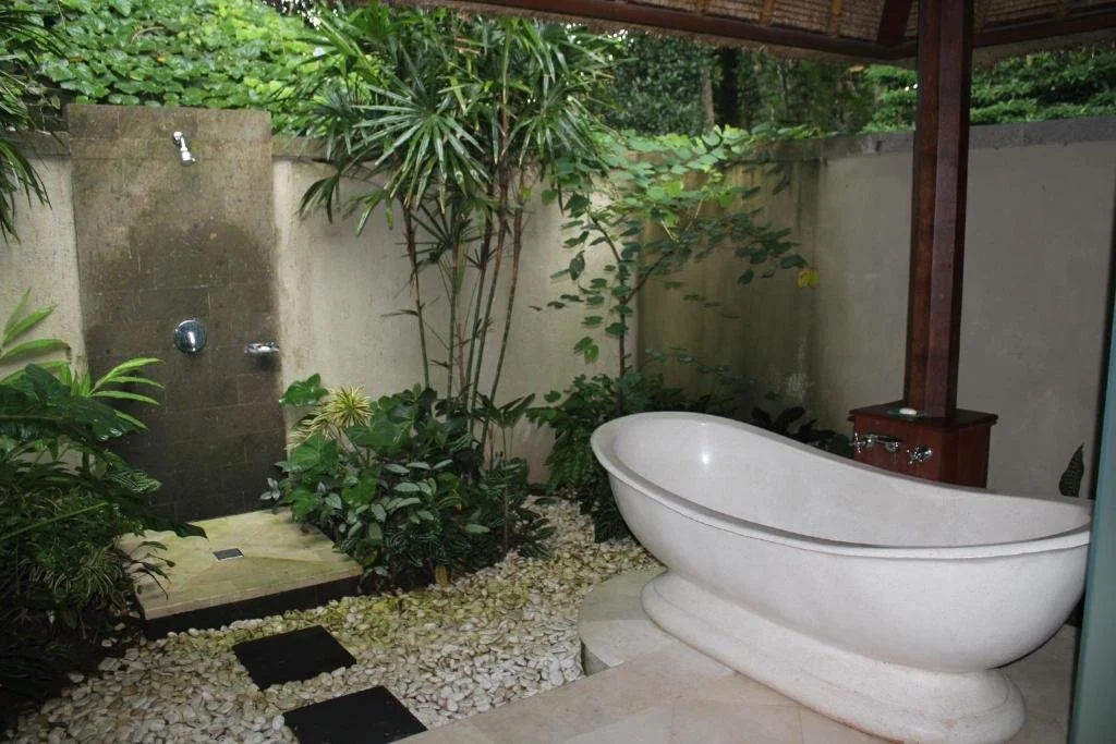 Outdoor bathroom with a white clawfoot bathtub, green plants, and a shower area with a stone platform and water faucet, enclosed by concrete walls and greenery.