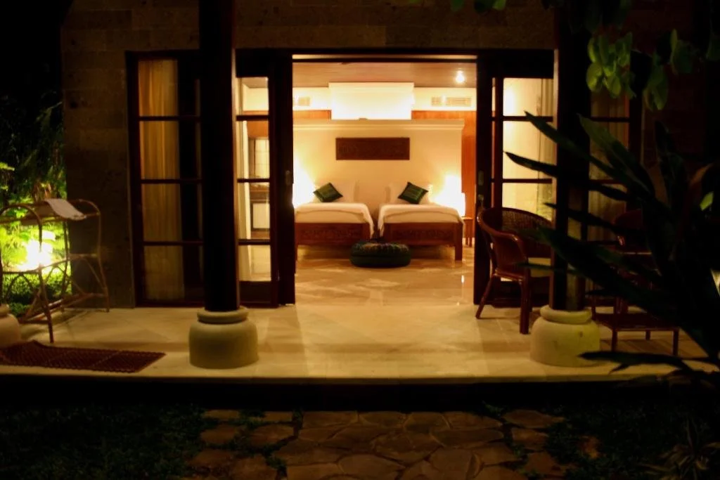 View of a hotel room with two beds, seen through open French doors, with warm lighting and wooden furniture.