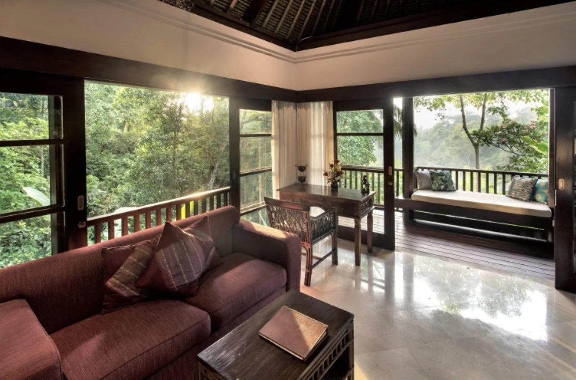 Living room with large windows overlooking a lush green forest, a burgundy sofa with pillows, a small wooden table with a book, a desk with a vase and flowers, and a swing bench with pillows on a wooden deck.