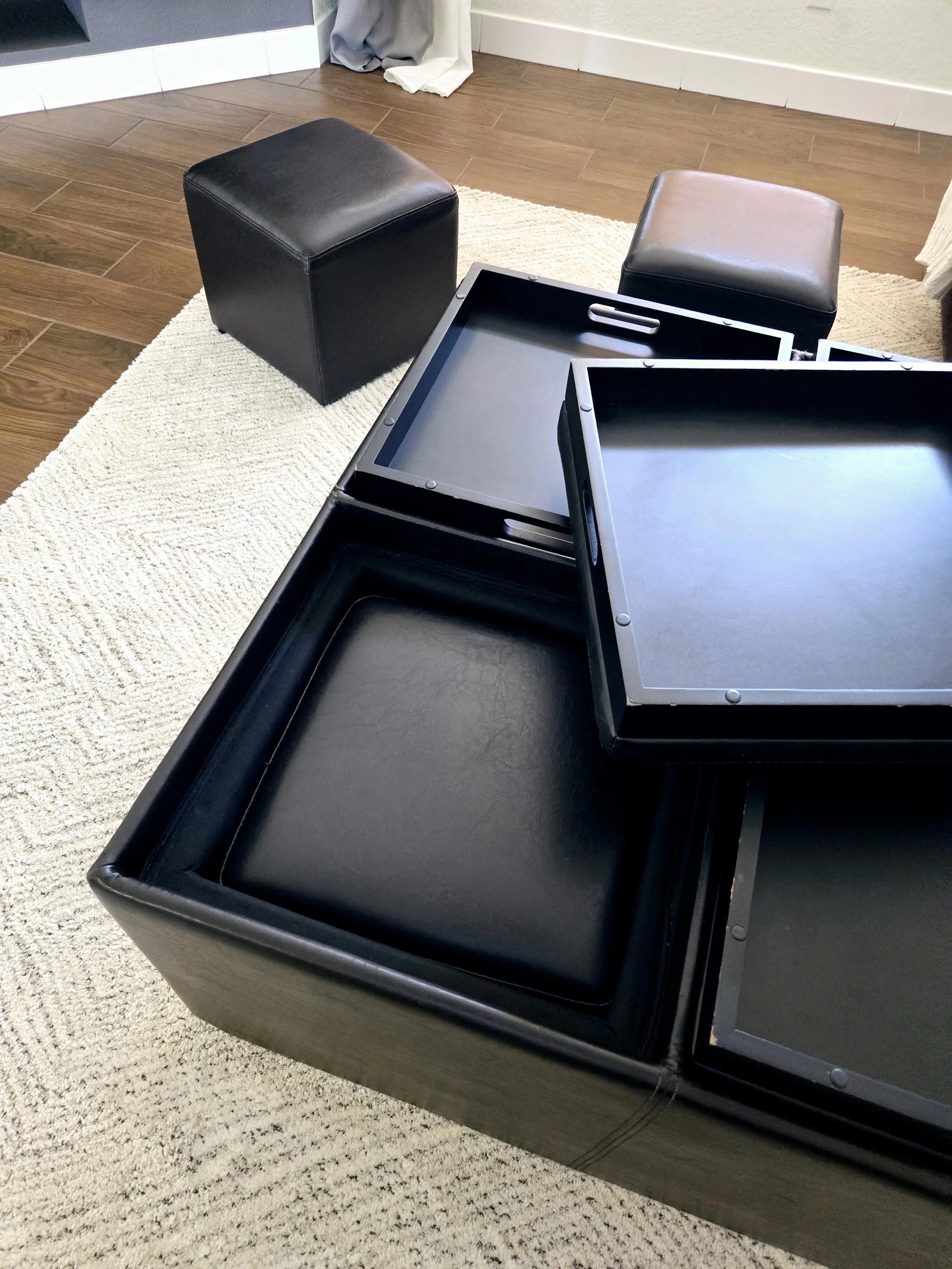 Black storage ottomans and trays on a beige carpet in a living room with hardwood floors, near a white wall and curtains.