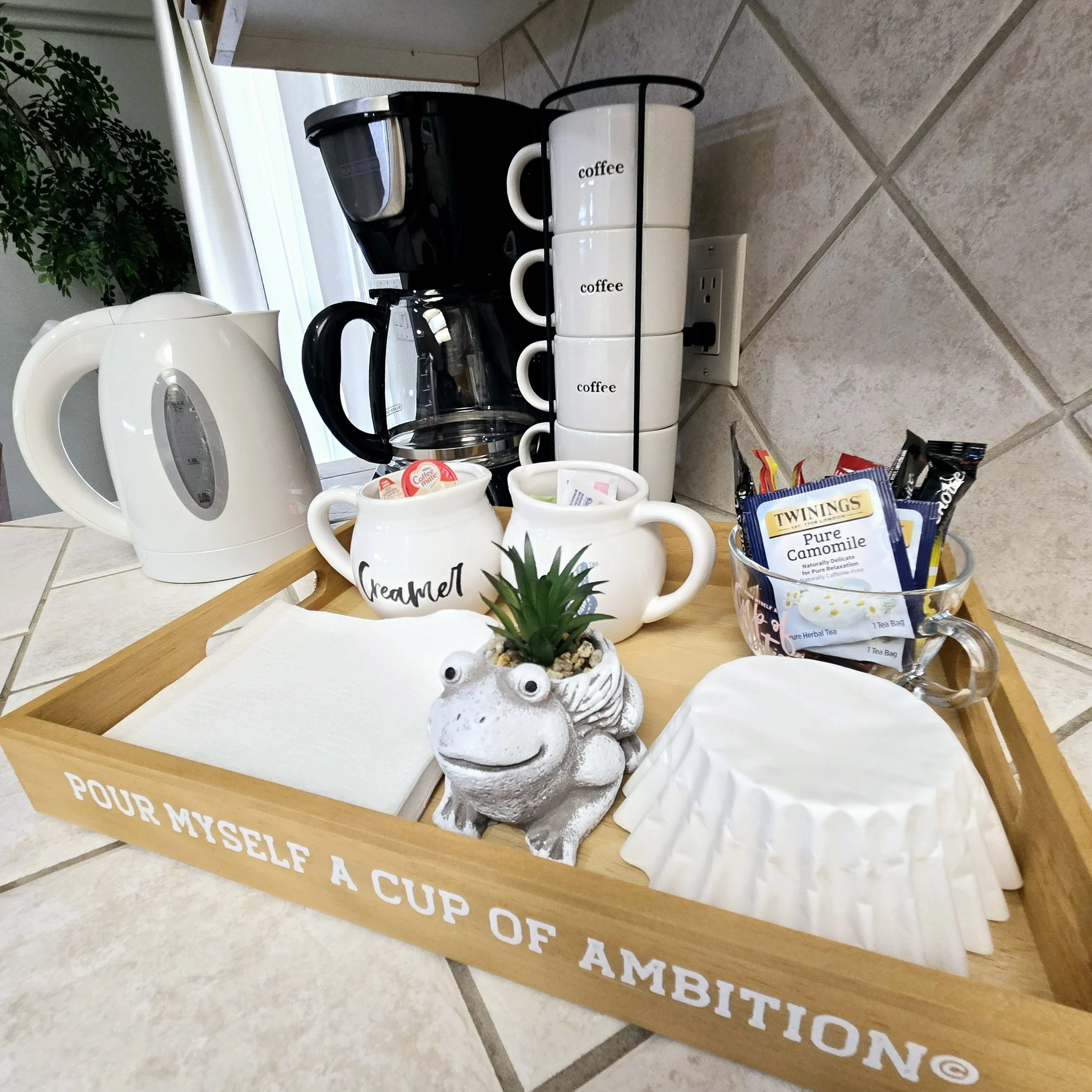 A kitchen countertop with a wooden tray containing a small potted succulent plant, white coffee mugs labeled "Coffee," a jar, a bowl of tea sachets, a white electric kettle, and a coffee maker, with a tiled wall and electrical outlets in the backgrou