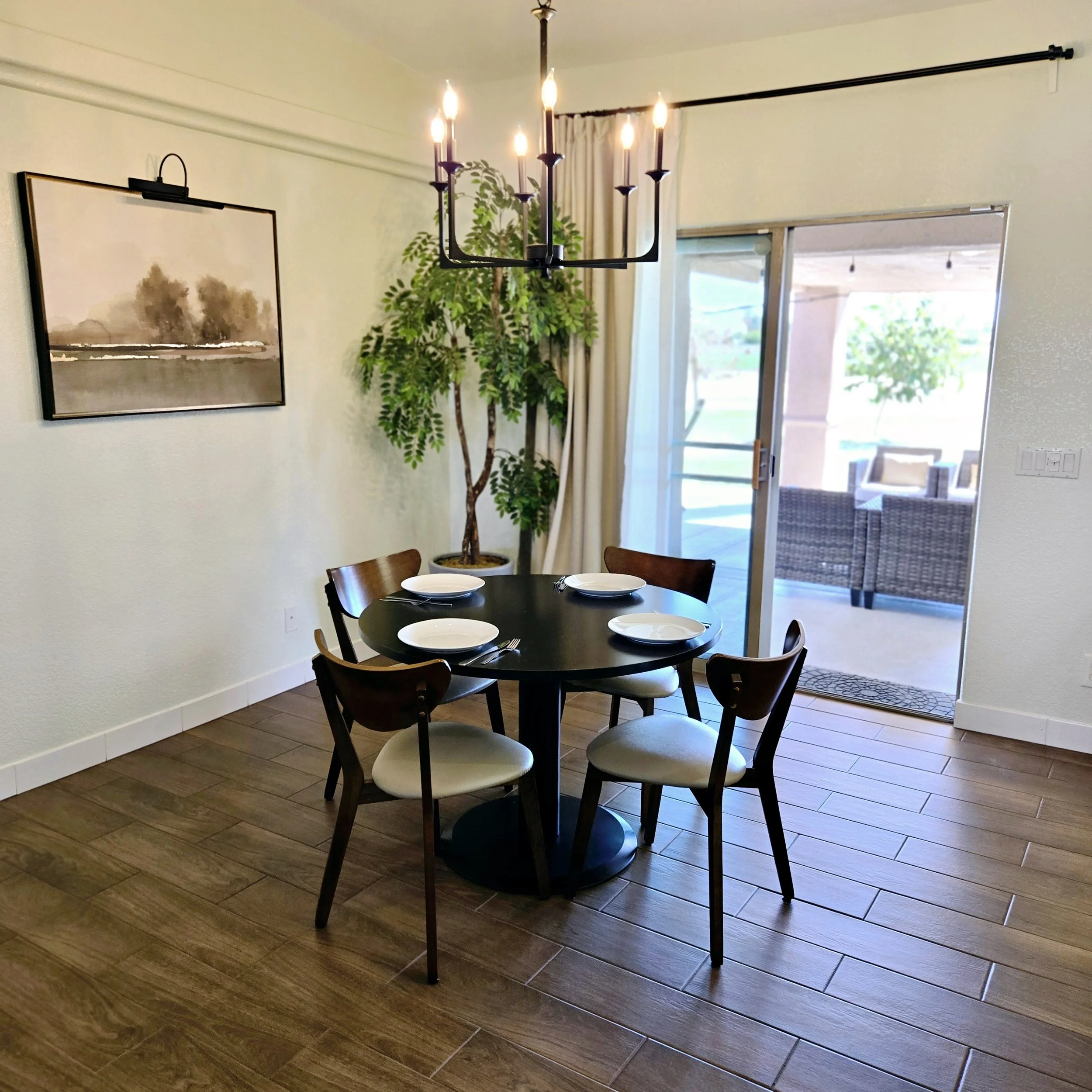 Dining area with a round black table set with four white plates and silverware, surrounded by four wooden chairs with white cushions. A chandelier hangs above, and there is a large potted plant in the corner near a sliding glass door leading to an ou