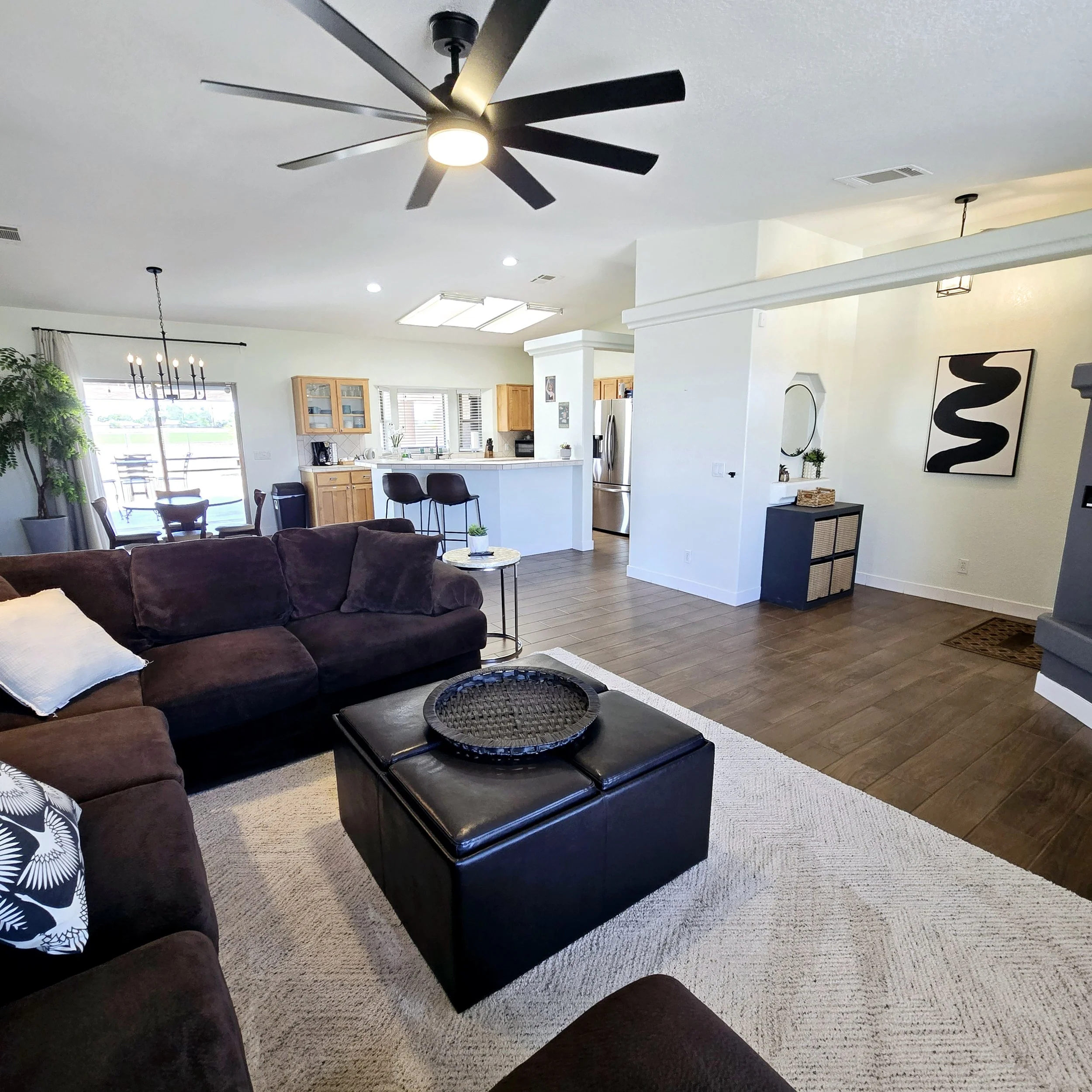 Living room with a dark brown sectional sofa, black ottoman with a tray, beige carpet, wood flooring, and an open kitchen with breakfast bar. Ceiling fan and modern art on the wall.