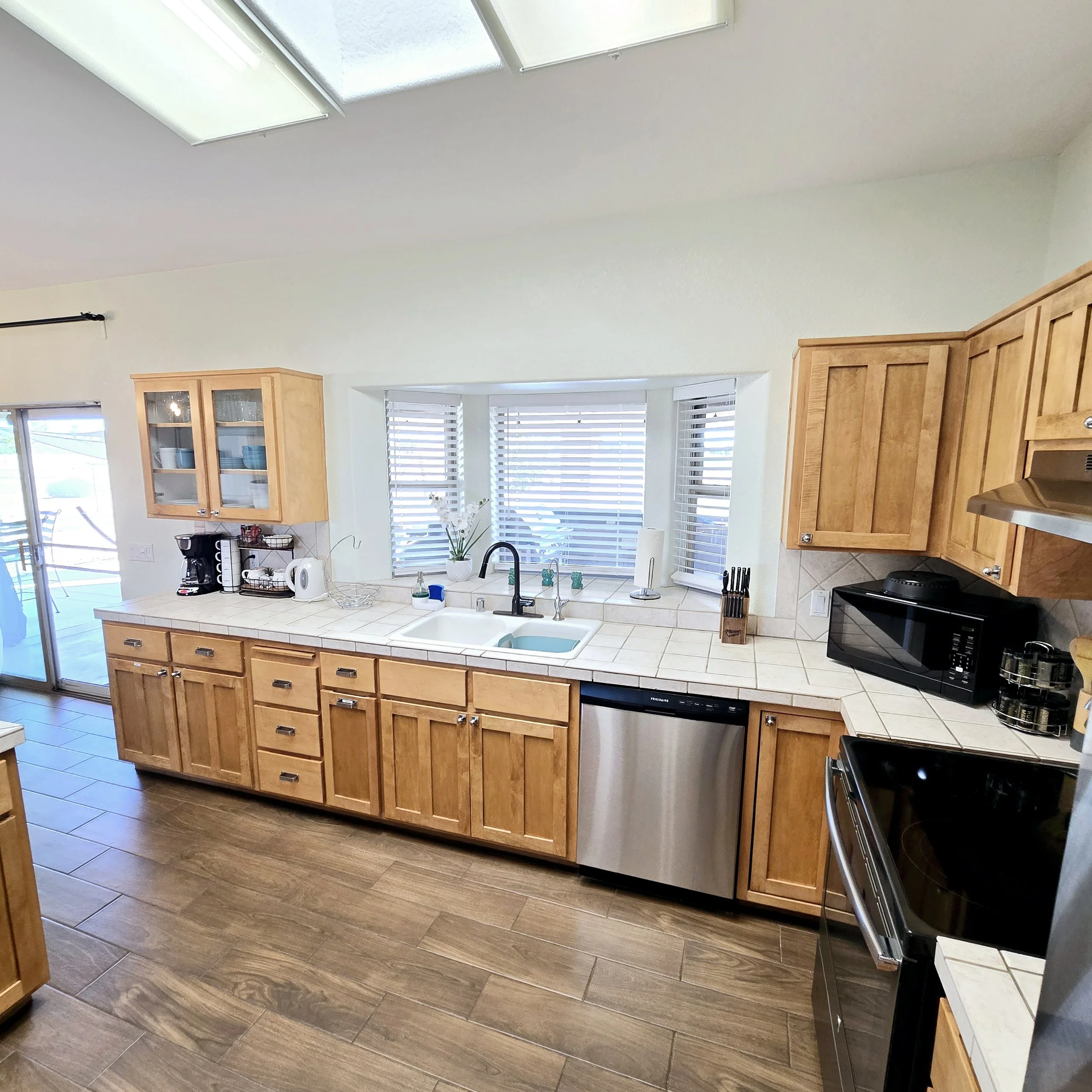 Kitchen with wooden cabinets, white tiled countertop, and stainless steel appliances. There is a double sink under a window, with a coffee maker, toaster, and microwave on the counters. A sliding glass door leads outside.