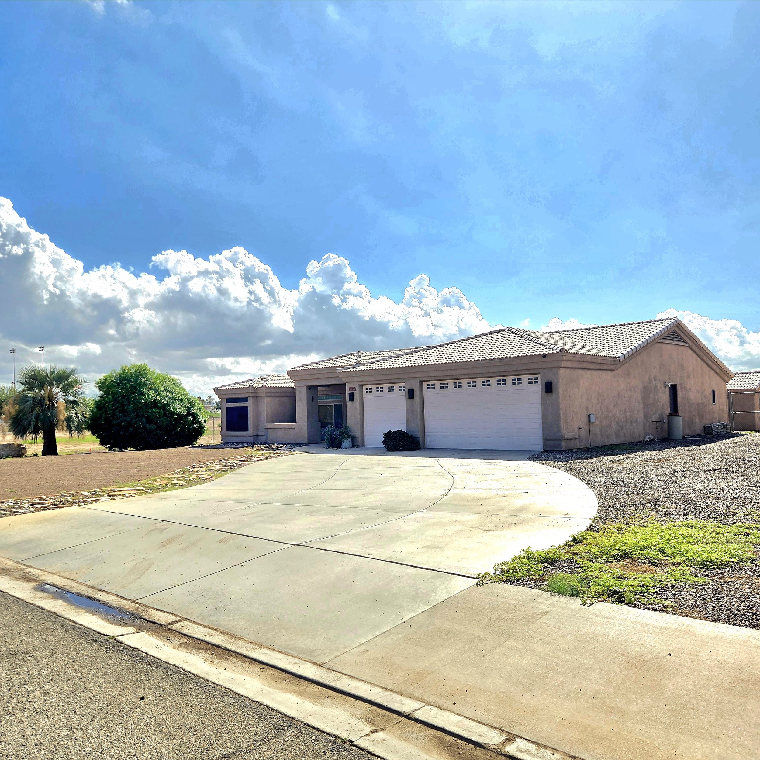 Single-story house with a beige exterior, a tiled roof, and a double garage. The house has a small front yard with some bushes and a gravel area. The driveway is curved and made of concrete, leading to the garage. The sky is partly cloudy with large 