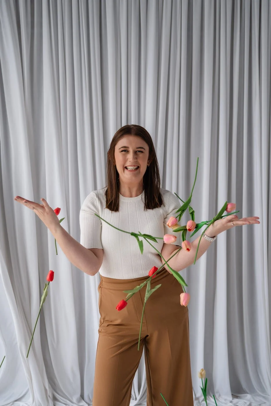Woman standing in front of a white curtain, holding her hands out and catching red and pink coloured tulip flowers