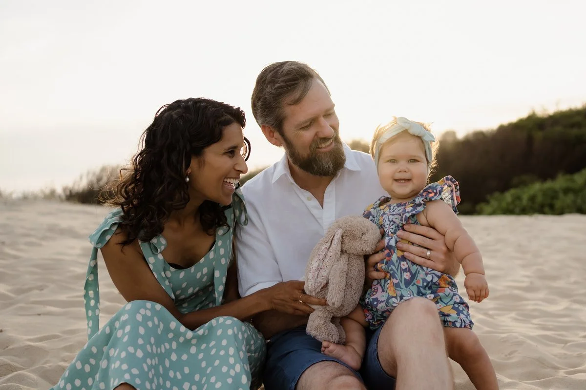 Toddler girl sitting on her fathers lap, with Mother sitting next to them at the beach in newcastle