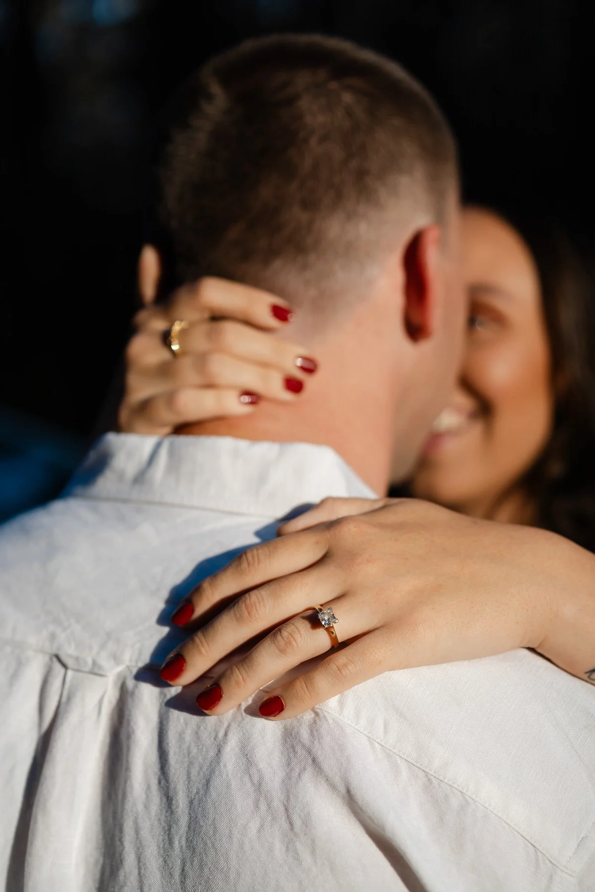 A couple embracing closely, with focus on the woman's hands showing an engagement ring on her finger, and her nails painted red.