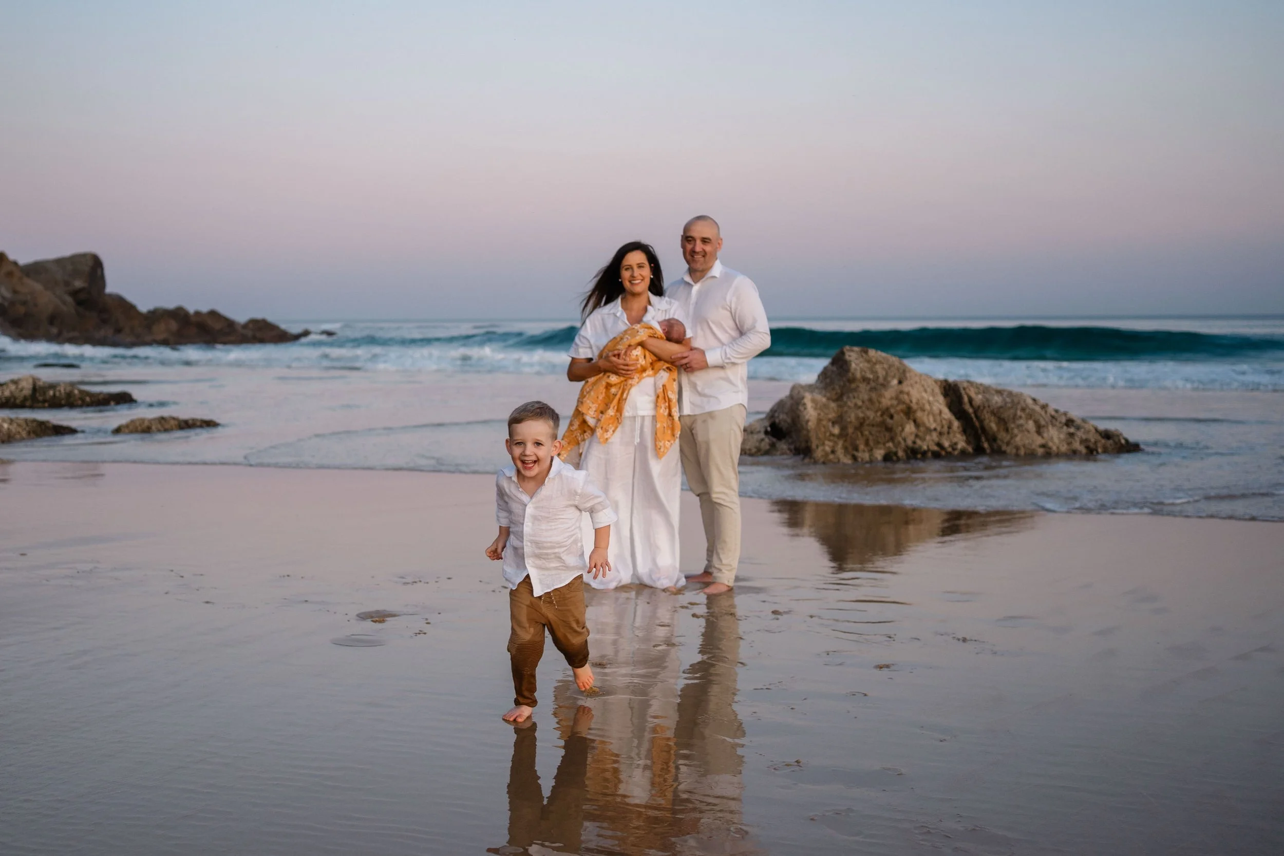 toddler running towards the camera at the beach, and parents stand in the background watching