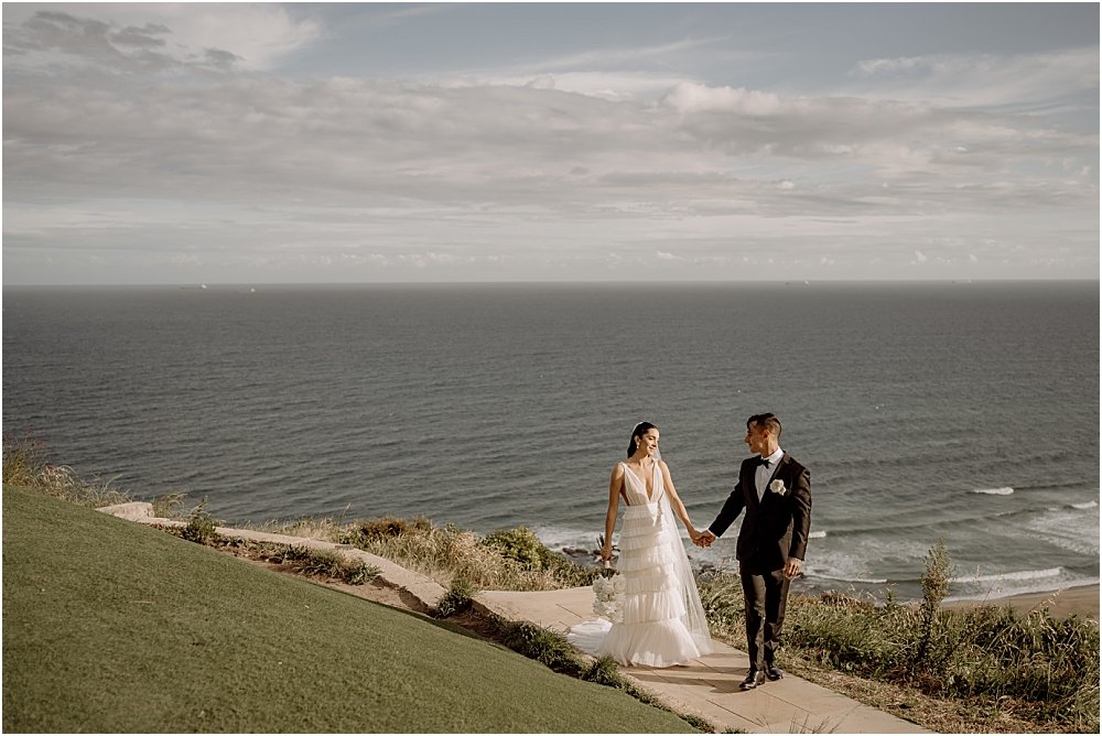 Groom holding brides hand and walking along path near lookout in Newcastle