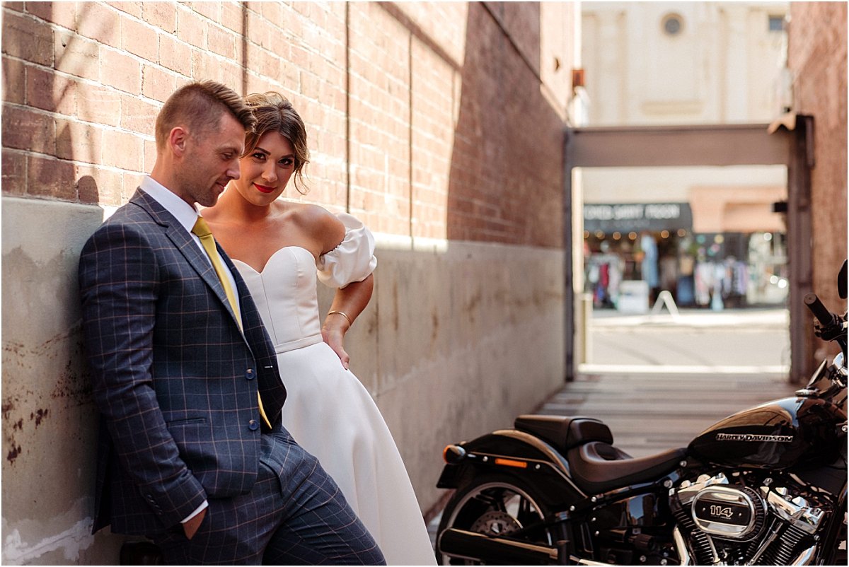 Bride and groom standing near black motorbike in brick alleyway