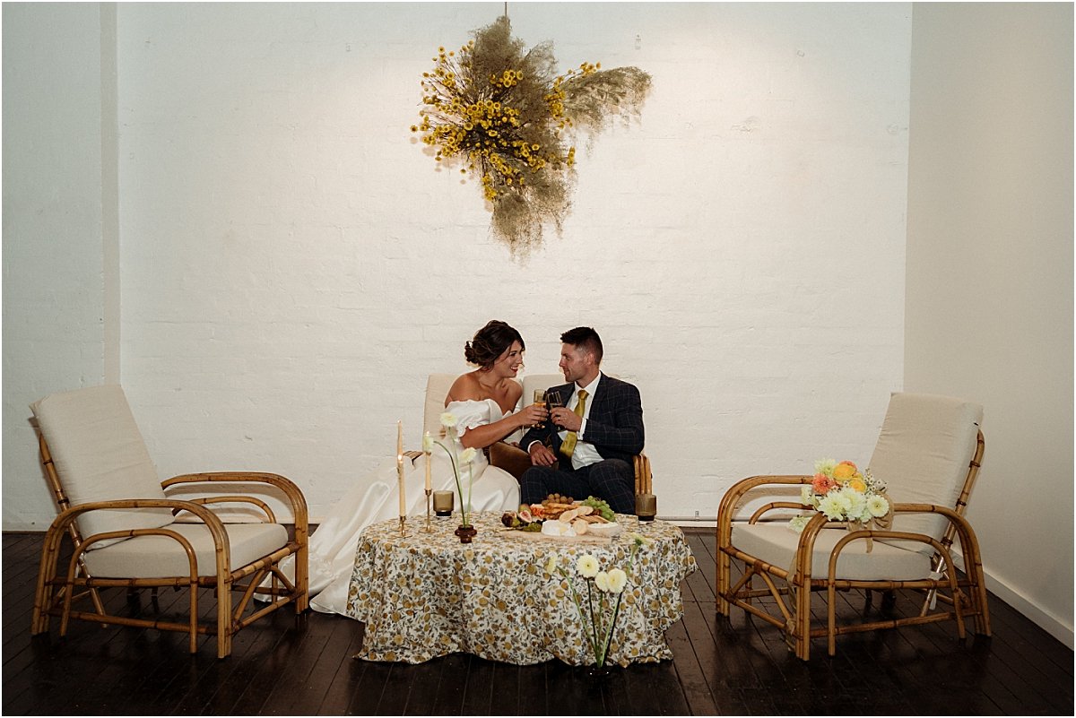 Bride and groom sitting on cane wedding furniture sharing grazing board together