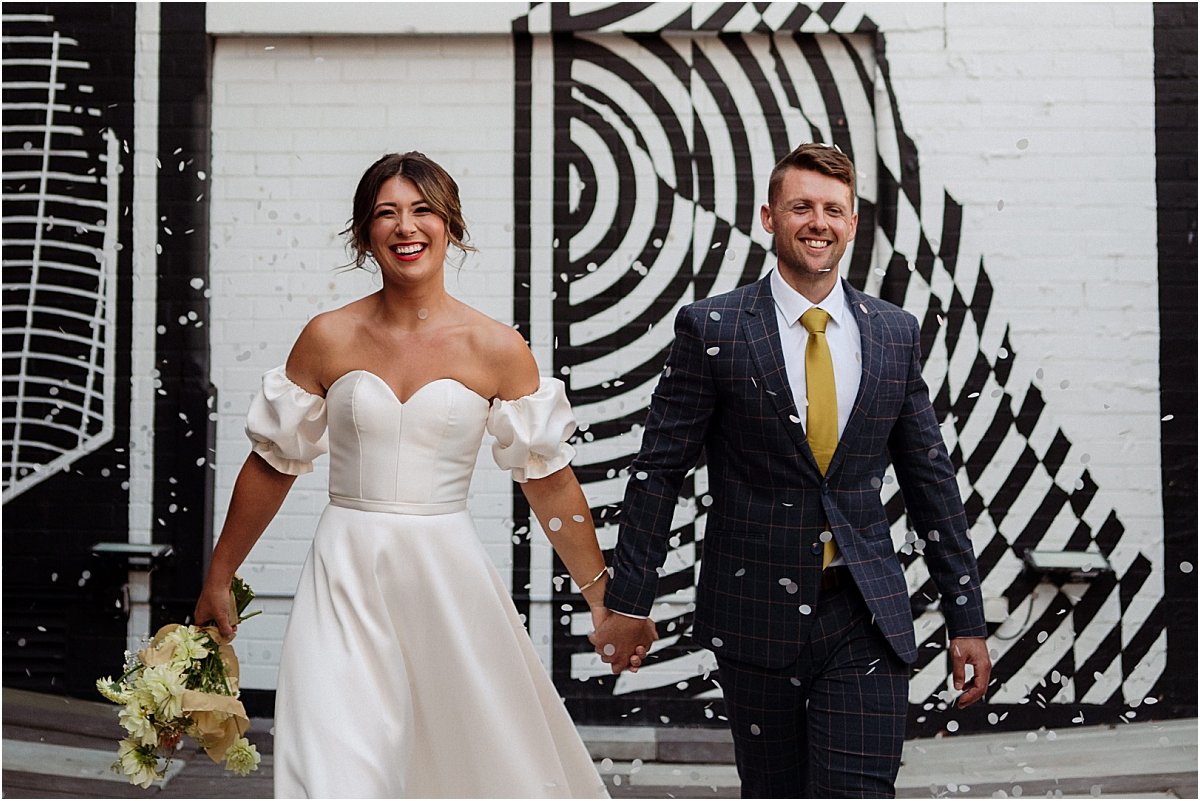 Bride and groom walking  into white bio-degradable confetti in front of black and white mural at the Owens Collective Islington NSW
