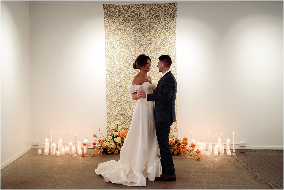 Bride and groom dancing together in front of retro fabric, pillar candles and orange and yellow floral nest