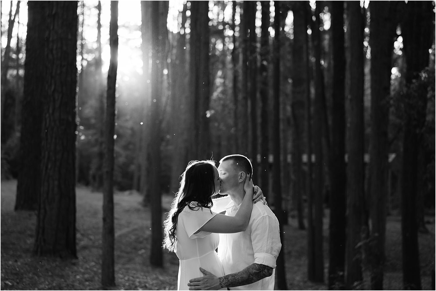 A black and white photo of a couple kissing in the woods.