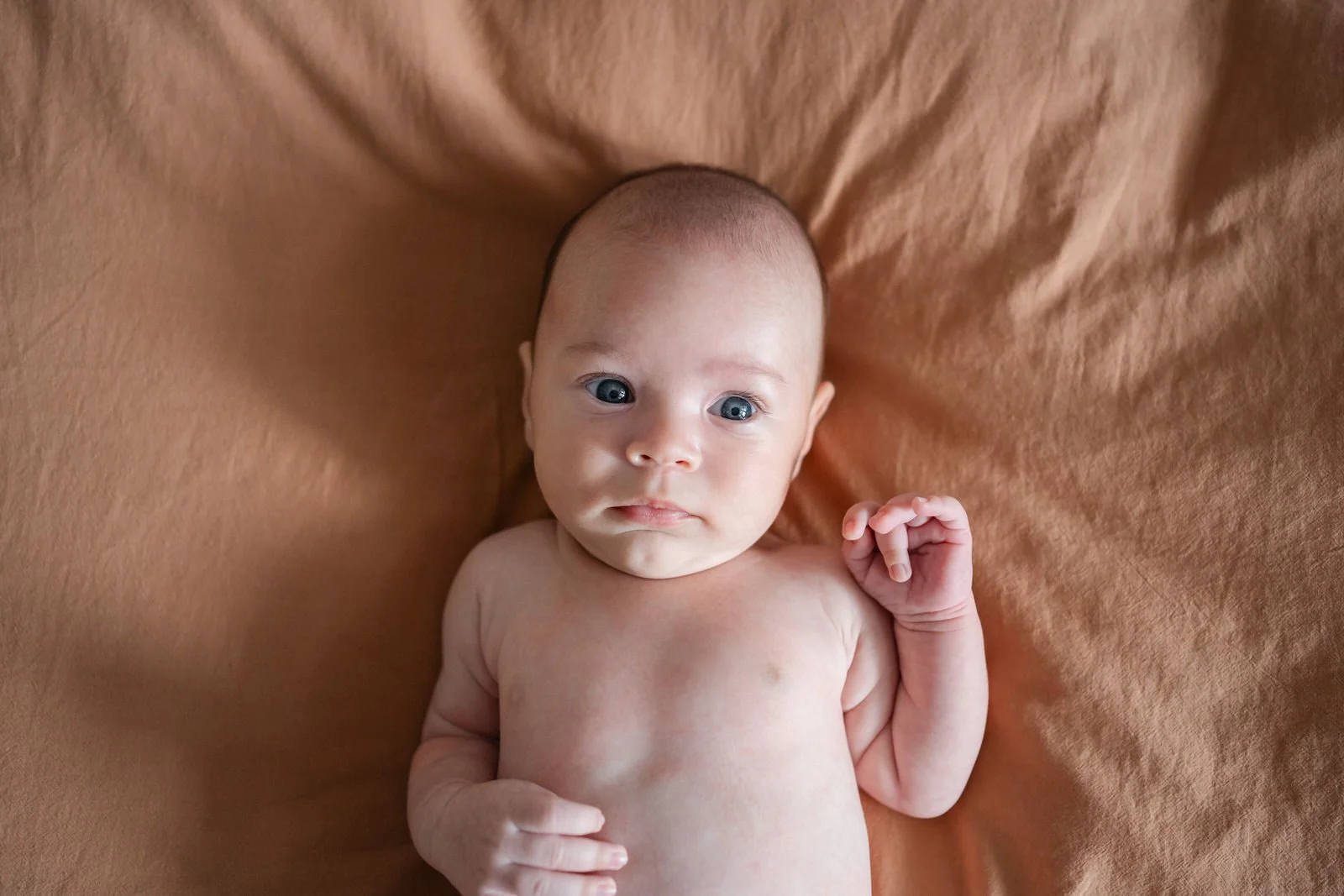 A newborn baby with blue eyes lying on a brown blanket, looking towards the camera with a curious expression.
