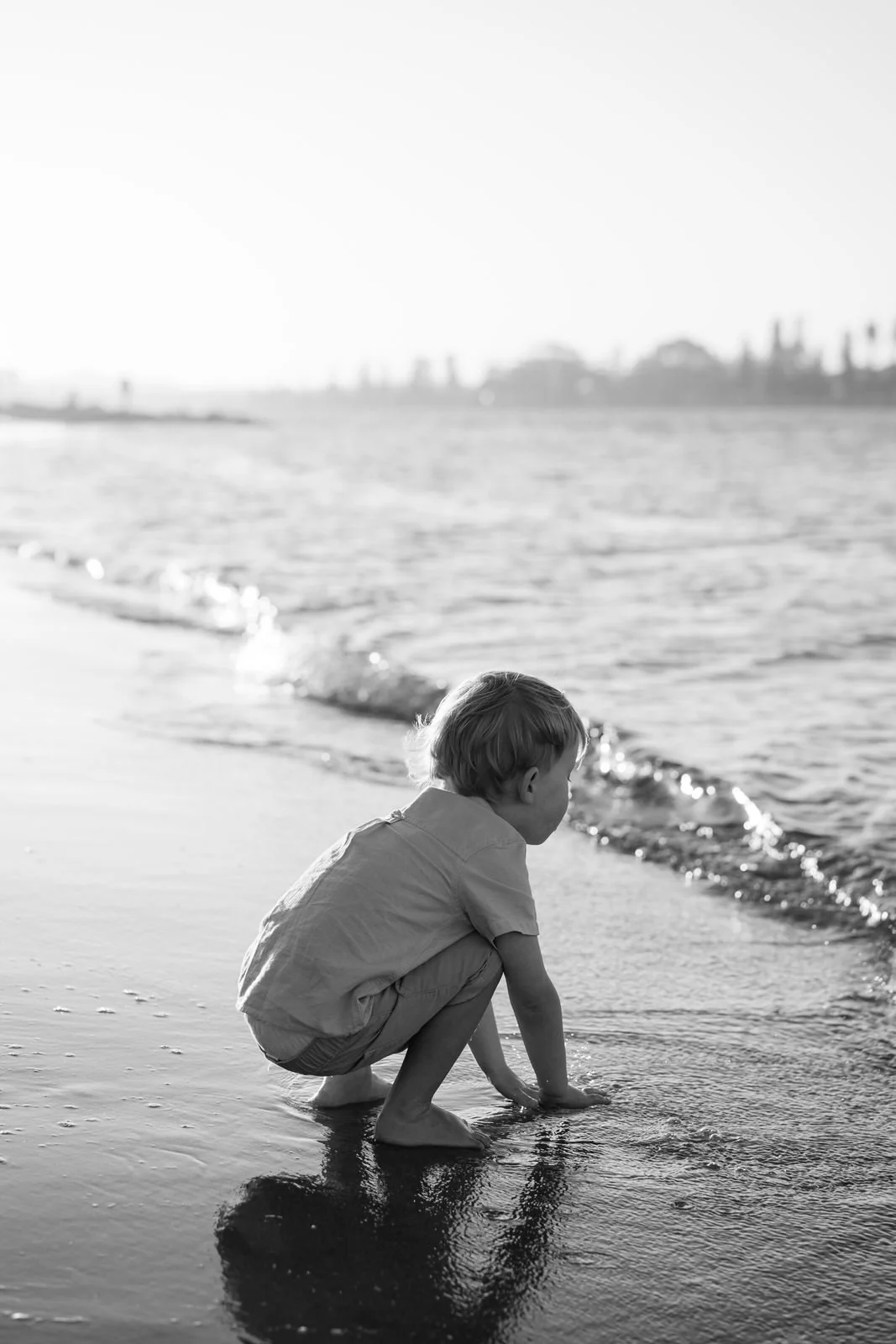 A young boy squats on the wet sand at the edge of the ocean, looking at the water, with city buildings in the distance on a sunny day.