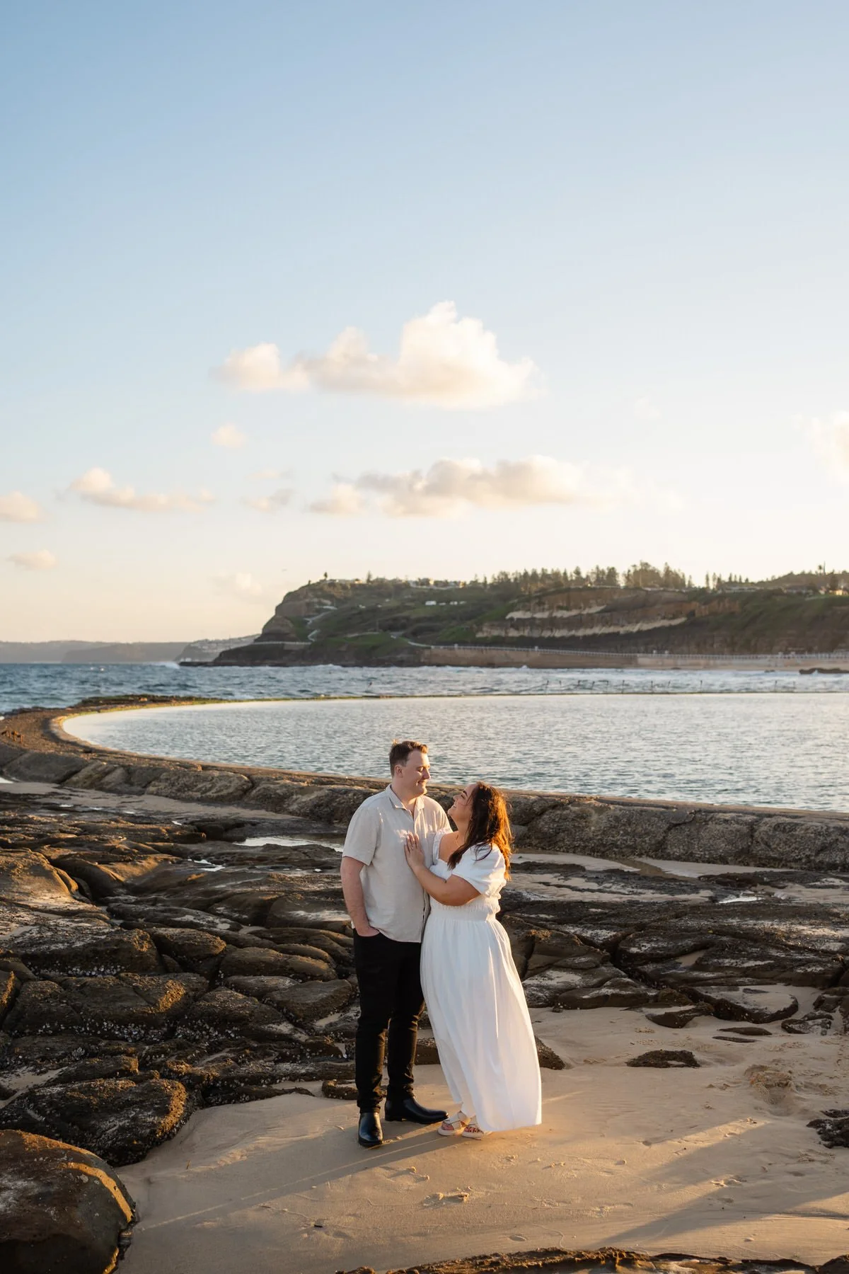 A couple standing on a rocky beach during sunset, with the ocean and hills in the background, looking at each other affectionately.