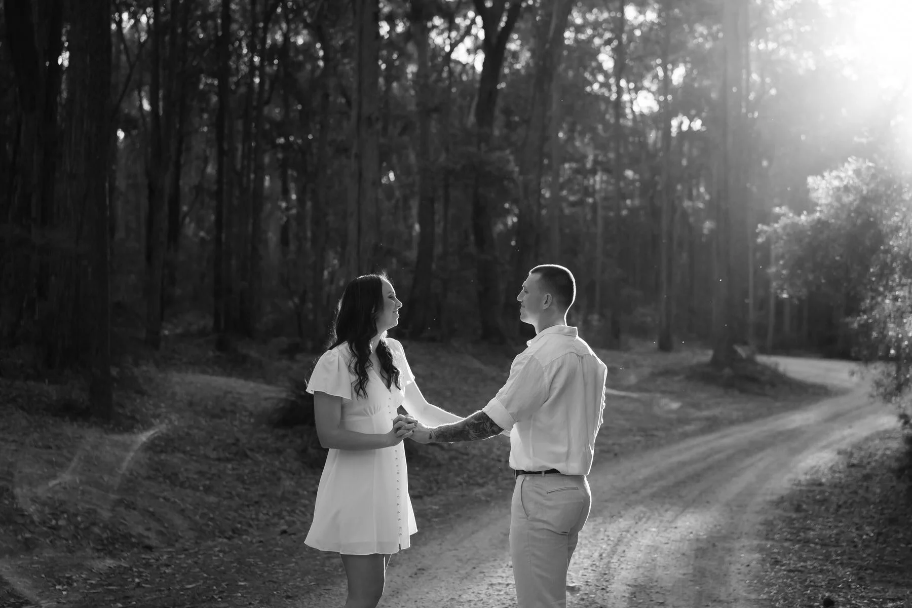 A black and white photo of a couple holding hands and looking at each other in a wooded outdoor area, with sunlight shining through the trees, for their engagement photoshoot in the Hunter Valley
