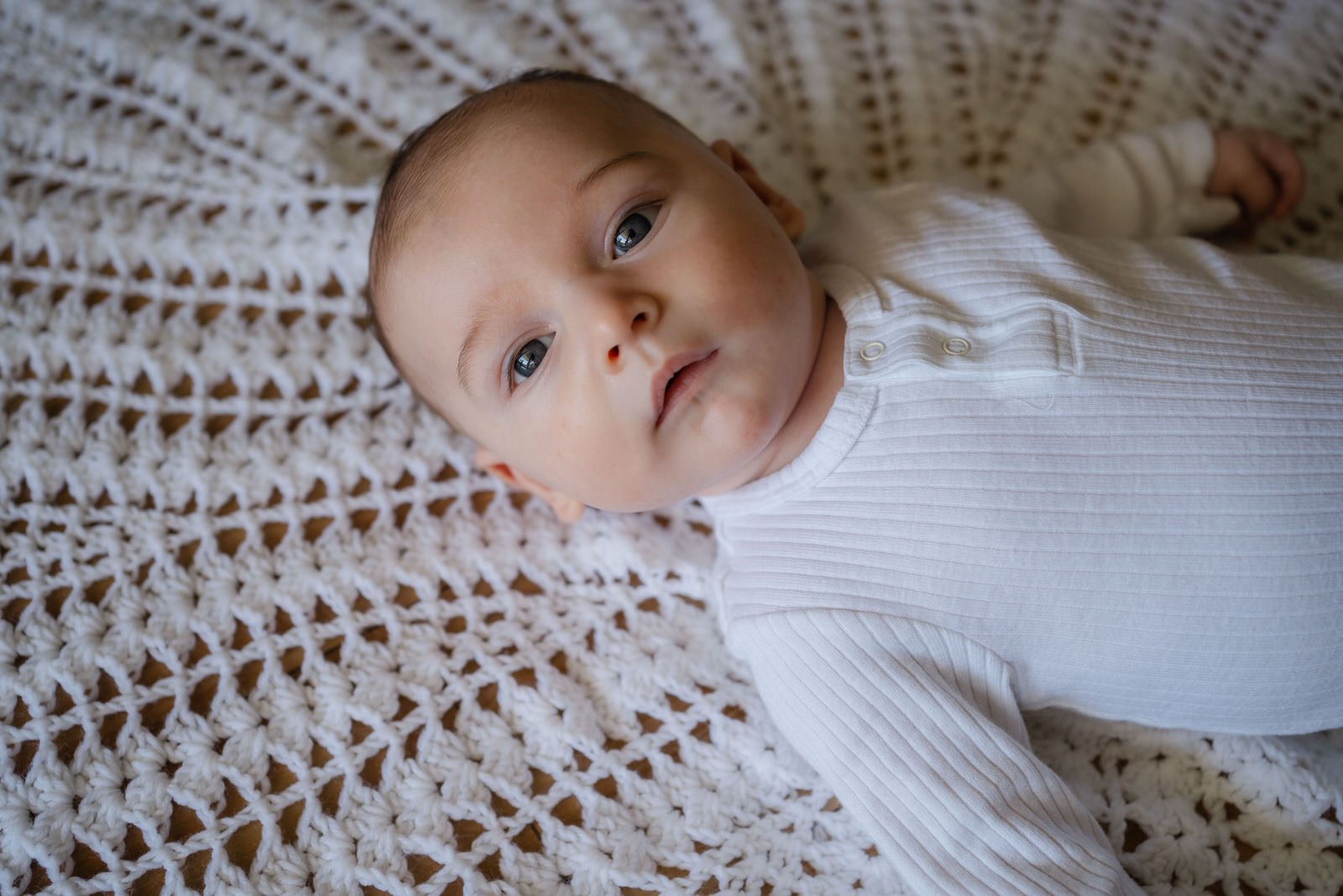 A baby with blue eyes lying on a textured white blanket, wearing a white long-sleeved ribbed shirt with buttons.