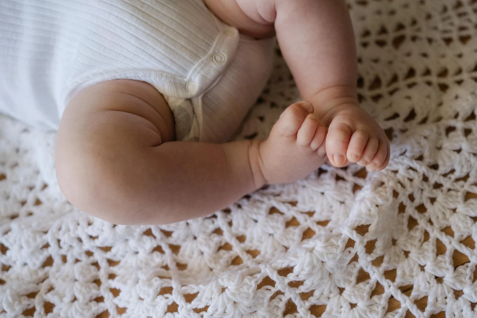 Close-up of a baby's arm, hand, and feet resting on a white, crochet blanket, wearing a light-colored onesie.