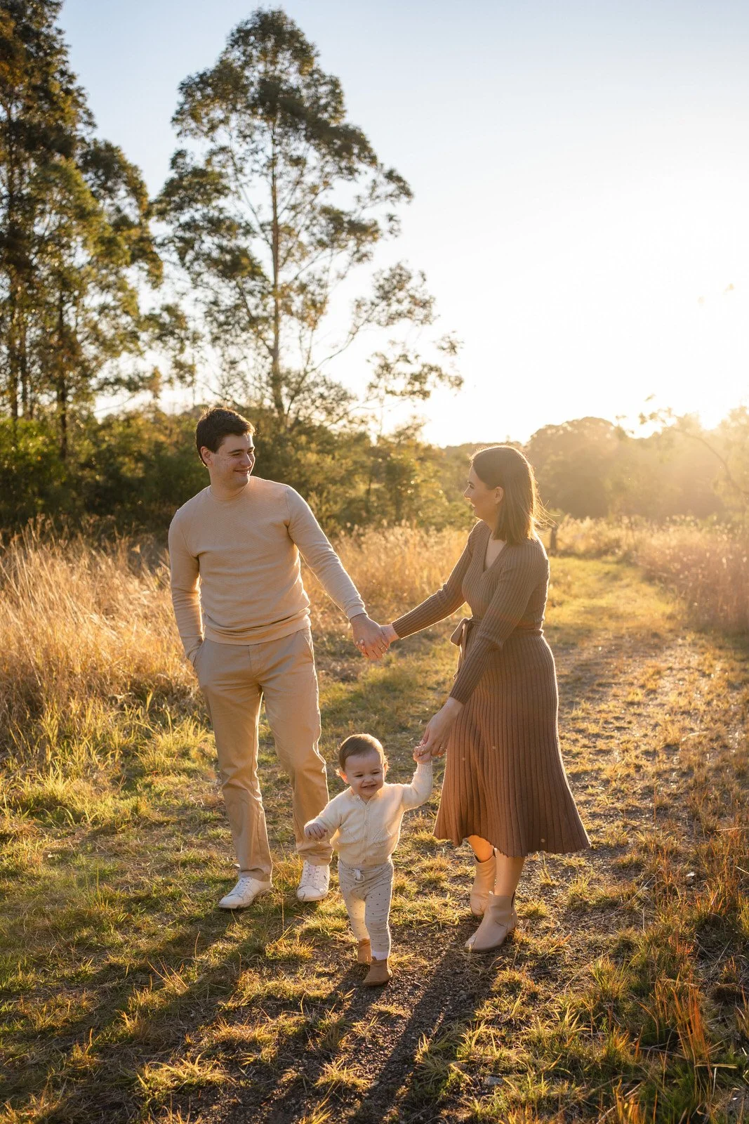 A family of three walking outdoors on a sunny day, holding hands, with trees in the background.