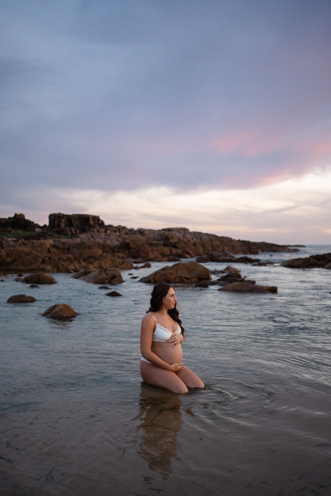 Pregnant woman kneeling in shallow ocean water at sunset, wearing white lingerie, with rocky shoreline and cloudy sky in background.