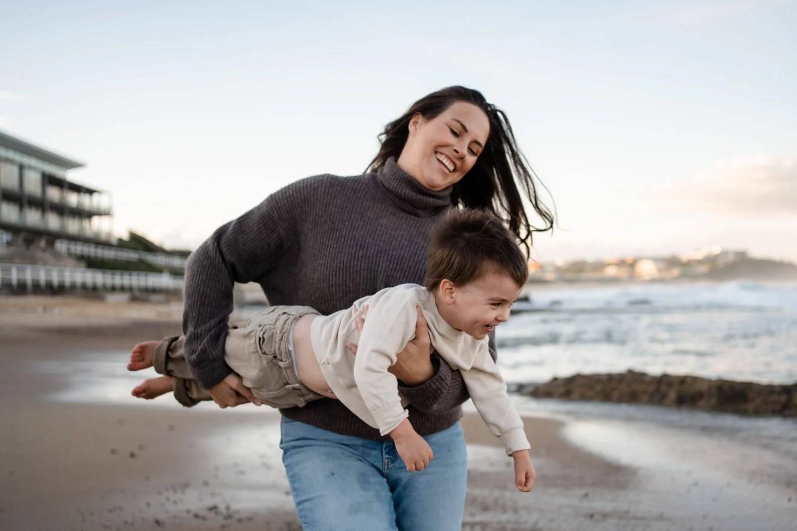 A woman playing with a young boy on the beach during the day, smiling and lifting him in her arms.