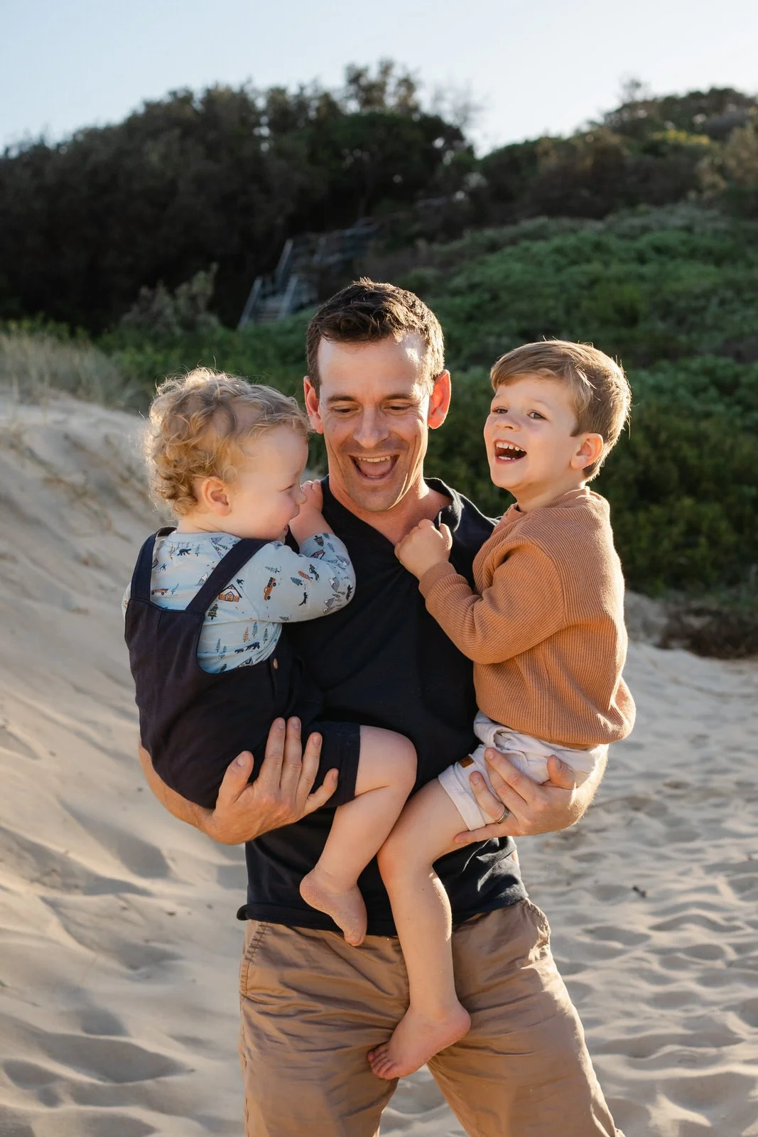 A man holding two young children on a beach, all laughing and smiling.