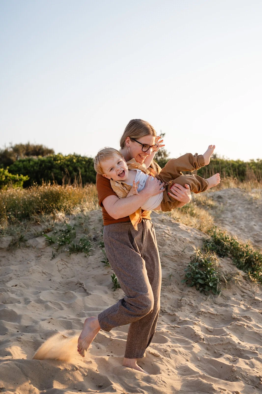 A woman with glasses and long hair playing with a young boy on a sandy beach during sunset, lifting him in the air and both laughing.