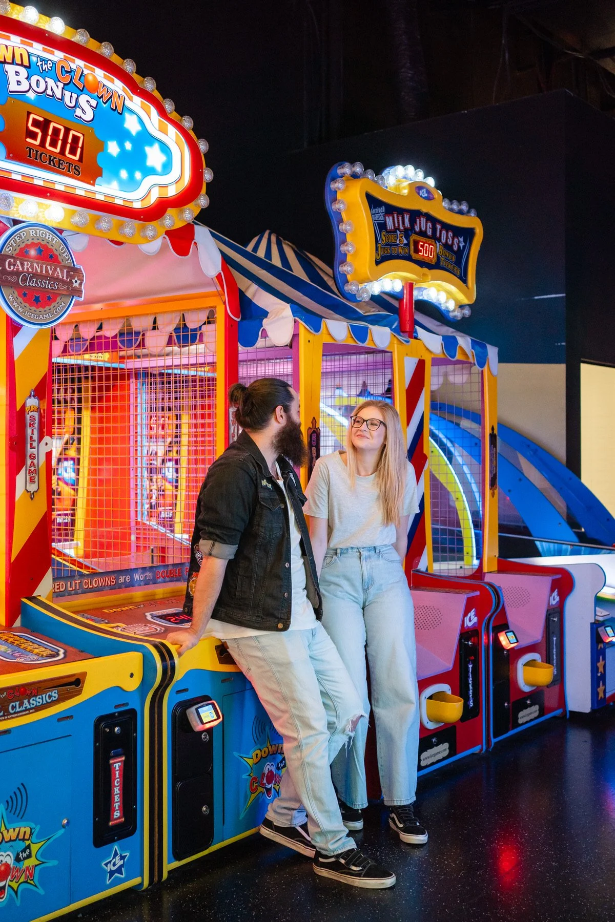 Two young people, a man with a beard and a woman with glasses, standing at an amusement arcade game surrounded by colorful dancing carnival machines and bright lights.