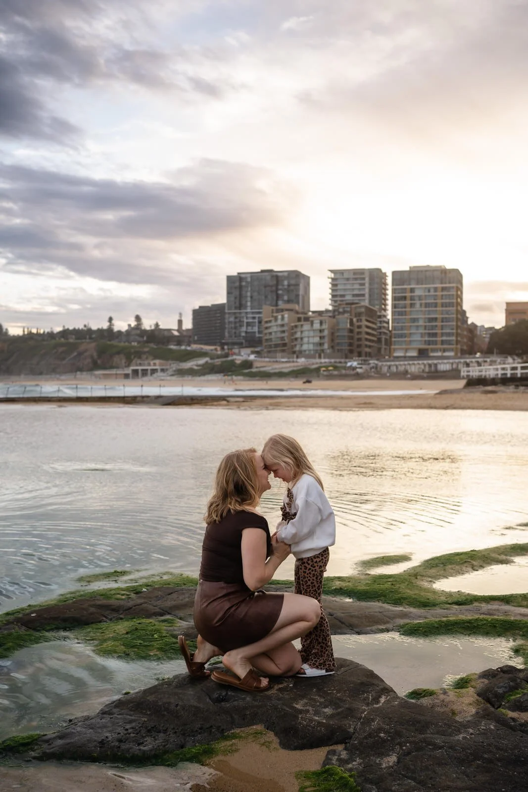 A woman and a young girl are touching foreheads and holding hands on rocks near a beach at sunset, with city buildings in the background.